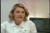 A woman with blonde hair in a white outfit sits indoors in front of a vintage computer monitor displaying green lines, reminiscent of the classic cyborg action seen in *Eliminators DVD (1986)*. She faces the camera with a neutral to slightly engaged expression, evoking the retro atmosphere of the film.