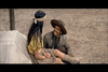 A man in a brown hat and jacket smiles up at a woman in a yellow shirt and beige skirt, who is sitting on a covered wagon. The woman has long black hair and a headband. Their outdoor setting on sandy ground evokes the timeless charm of classic western scenes, reminiscent of those featured in Rio Conchos DVD (1964).