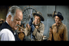 Three men in Western attire stand indoors. The man in front holds a cigar, the middle man wears gloves and holds metal shackles, and the third man looks on. A large wooden wagon wheel hangs on the wall behind them—an evocative scene reminiscent of those found in Rio Conchos DVD (1964), a classic western highly prized by collectors seeking rare movies on DVD.