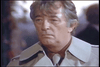 A man with gray hair wearing a light-colored trench coat stands indoors, looking serious and focused. The blurred background suggests other people or activity behind him, reminiscent of a scene from the rare classic movie “The Amsterdam Kill DVD (1977).”.