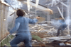 Two people in blue uniforms stand amidst a chaotic, debris-strewn scene, one aiming a gun at the other as smoke swirls around them. Shattered wood and metal litter the area, capturing the gritty intensity of The Amsterdam Kill DVD (1977).