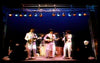 Four musicians in white suits perform on a brightly lit stage at night. Three play guitars at the front while a drummer is visible behind them. Stage lights and metal rigging frame the energetic scene, reminiscent of Lennon/McCartney classics featured on the Beatlemania DVD (1981).