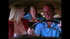 Three people sit in a car: a blonde woman in a red-patterned top evocative of the 1991 classic film “Whore,” now available on DVD, a man driving and laughing, and another man in the backseat, all sporting blue shirts. The moment is lively and comedic.