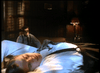A man in a white shirt lies on a bed, eyes closed, as another man in a suit—evoking classic mafia style—sits shadowed in the background of a dimly lit room. Scene inspired by Killer Instinct DVD (1992).