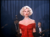 A woman with short curly blonde hair and an off-shoulder red dress, like a character from "Killer Instinct" (1992) DVD, stands before a dark blue curtain as musicians in tuxedos play in the background.