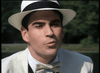 A man in a white suit, bow tie, and fedora stands outdoors by water, channeling the classic mafia style seen in "Killer Instinct" (1992), now available on DVD.