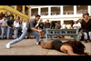 A crowd watches as one person stands over another lying face-down on the ground in an outdoor courtyard, evoking a dramatic moment from a martial arts film. In the background, a table and building add to the cinematic setting—perfect for fans seeking rare movies or collectors searching for that elusive Only the Strong (1993) DVD.