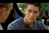 A young man with short dark hair and a serious expression listens intently to someone offscreen in a scene from Only the Strong (1993) DVD. He wears a blue shirt, and the background is blurred as a figure passes by, capturing the focused atmosphere characteristic of martial arts films.