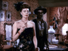 A woman in a strapless dark dress holds a drink in an ornately decorated vintage room, reminiscent of scenes from old films. A man in a hat and vest stands behind her—similar to moments in "Outlaw Women (1952) DVD.