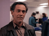 A middle-aged man with curly dark hair, in a plaid shirt and brown leather jacket, stands indoors. The background is an office where people discuss rare films like "A Deadly Silence (1989) DVD.