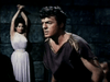 A man in ancient clothing stands tensely as a woman in a white dress is tied to a stone wall—a dramatic scene from the rare, hard-to-find vintage classic "The Minotaur (1960) DVD.