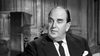 A middle-aged man with a receding hairline in a suit and tie sits in a wooden chair against a paneled wall, looking serious—a scene evoking the classic British mystery, Murder at the Gallop (1963) DVD.