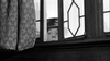 A person in a hat peers through a decorative window beside a patterned curtain, only their eyes and part of their face visible, evoking the black-and-white style of "Murder at the Gallop (1963)" DVD.