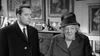 A suited man stands beside an older woman in a hat and coat, indoors with framed paintings and décor behind them. This black-and-white image from the Murder at the Gallop (1963) DVD evokes a classic British mystery atmosphere.