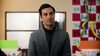 A man with dark hair in a gray suit jacket stands in an office with colorful streamers and a big calendar, appearing to be listening or reacting as if discussing the Game of Love (2023) DVD.