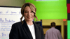A woman with light brown hair in a black blazer and white shirt stands smiling by a whiteboard and green walls, resembling a romantic scene from the "Game of Love (2023) DVD." A blurred person appears in the background.