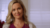 A woman with wavy blonde hair and a gentle smile, dressed in a purple top and light gray sweater, stands indoors by the blinds—capturing the nostalgic charm of "Hearts of Christmas (2016) DVD.