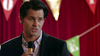 A man in a dark suit holds a microphone, standing before red curtains and pennant banners, evoking the vintage charm of films like "Hearts of Christmas (2016)" on DVD.