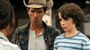 A man in a cowboy hat and plaid shirt stands beside a curly-haired boy in a blue striped t-shirt, both outdoors—reminiscent of scenes from "The Real West (2024) DVD," capturing the romance and adventure of the American frontier.