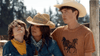 A woman in a straw cowboy hat hugs two boys—one in a yellow bandana, the other in a white hat and horse-print shirt—outdoors with trees behind them, echoing the frontier spirit of "The Real West (2024) DVD.