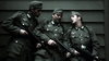 Three men in WWII German uniforms stand together, rifles in hand, deep in conversation against a dark wooden wall—a tense moment captured from Spoils of War (2009) DVD.