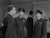 Three men in military uniforms converse indoors in this black-and-white scene from The Dam Busters (1955) DVD. Two face the camera, one faces away; a coat and hats hang behind them, with a fireplace to the right.
