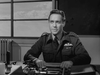 A man in a military uniform with pilot wings sits at a desk, facing the camera. On his desk is The Dam Busters (1955) DVD, emphasizing British war films. A window in the well-lit room shows a partly cloudy sky.