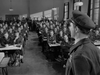 A uniformed officer addresses a large group of airmen in a classroom, evoking memorable scenes from the British war film The Dam Busters (1955) DVD, as sunlight streams through tall windows and everyone listens attentively.