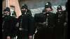 A group of uniformed police officers in tall helmets and dark coats stand outside a brick building, evoking scenes from the "Ned Kelly (1970) DVD.