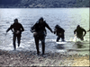 Four people in dark wetsuits and masks emerge from the water onto a rocky shore, carrying gear—reminiscent of scenes from classic war films like Submarine X-1 (1968), now available on DVD.