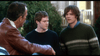 Three men talk outside; one wears a brown leather jacket, facing two younger men in dark sweaters. Trees and a building set the scene—like Thomas Kinkade’s Christmas Cottage (2008) DVD brought to life.