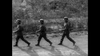 Three soldiers in uniform and helmets march in a line on a dirt road, their shadows stretching out behind them. Trees and bushes fill the background in this dramatic black-and-white image from Silent Raiders (1954) DVD.
