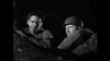 Two men in military uniforms sit together at night, partly hidden by a tree or camouflage. This black-and-white scene captures the classic World War II atmosphere of Silent Raiders (1954) DVD. One wears a helmet; the other does not.