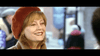 A woman in a red beret and tan coat smiles warmly outdoors, evoking the cozy holiday spirit of "Noel (2004)" DVD, with festive buildings and blurred people in the background.