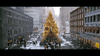 A large, decorated Christmas tree with bright lights stands in a city plaza, just like scenes from Noel (2004) DVD. People gather as snow falls softly, surrounded by tall buildings and shops in this festive holiday setting.