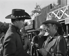 A sheriff in a wide-brimmed hat gives a revolver to a cowboy on the street of a Western town, capturing classic film spirit—scene from "Gun Duel at Durango (1957)" DVD, with wooden buildings and townsfolk in the background.