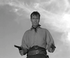 A man in vintage Western attire stands outdoors with two pistols drawn, evoking classic films. This black-and-white scene is from the "Gun Duel at Durango (1957) DVD," with wispy clouds in the sky behind him.