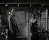 A man and a woman stand on a porch at night, evoking classic film scenes; he looks left, serious, while she watches intently—capturing the suspense of "Gun Duel at Durango" (1957) now available on DVD.