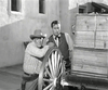 Two men in western attire stand by a wagon wheel and crates in a rustic setting, as seen on the black-and-white "Gun Duel at Durango (1957) DVD," which captures the essence of rare and classic Western movies.