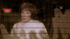 An older woman with short brown hair and a white top gazes through a night window, her reflection mixing with city lights—capturing the quiet longing seen in rare DVDs like A Heavenly Christmas (2016) DVD.