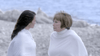 Two women wrapped in white blankets stand together on a tranquil beach, echoing the warmth and wonder found in "A Heavenly Christmas (2016) DVD.