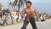 A muscular, shirtless man wielding a sword stands defensively on a Panama beach as onlookers watch—just like an iconic scene from Morgan The Pirate DVD (1960), evoking classic Steve Reeves pirate adventures.