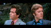 Two men in suits sit outdoors, each looking opposite ways, recalling the classic heist vibe of The Hot Rock (1972) DVD. One wears a striped jacket and pink shirt; the other has a blue shirt with tie and gray jacket.