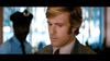 A serious-looking blond man in a suit and tie stands indoors, channeling Robert Redford’s style from the film "The Hot Rock," as a blurred police officer appears behind him. Now available: The Hot Rock (1972) DVD.