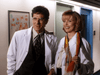 Two doctors in white lab coats smile in a hallway with framed art. The man has dark hair and glasses; the woman wears a colorful scarf. Their camaraderie mirrors heartfelt moments from the "Color Me Perfect (1996) DVD.