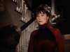 A woman with dark hair and red earrings in a high-necked red velvet top stands indoors near a staircase, reminiscent of a scene from the rare "Color Me Perfect (1996) DVD" about an intellectually disabled woman.