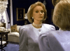 A woman with short blonde hair in a lavender blazer and pearl choker admires her reflection in a dim room decorated with art, a chandelier, and shelves featuring classic movies like "People Like Us (1990)" on DVD.