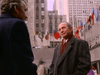 Two men in suits talk outside at Rockefeller Center, NYC, with flags and tall buildings behind them—like a scene from “People Like Us (1990)” on DVD. One faces the camera; the other is seen from behind.