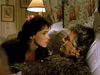 A woman leans toward an older man lying in bed, both sharing a moment in a softly lit room decorated with floral curtains and pictures—evoking the warmth of classic films like "People Like Us (1990)" on DVD.