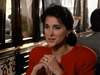 A woman with dark hair in a red outfit and earrings sits indoors at a table, hands clasped and attentive, amid ornate mirrors and decor—a scene evoking classic moments from the "People Like Us (1990) DVD".